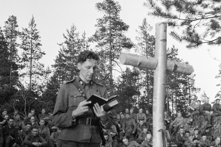 sa-kuva, kenttähartaustilaisuudesta, pappi ristin äärellä lukemassa, takana sotilaat maastossa kuuntelemassa