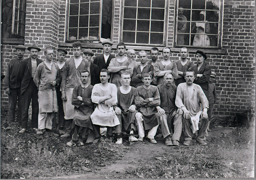 Kalliokosken lasitehtaan työntekijöitä vuonna 1922 käyttöön otetun tehdasrakennuksen seinustalla. 
Kuva: Miehikkälän kotiseutumuseon kokoelmat. 